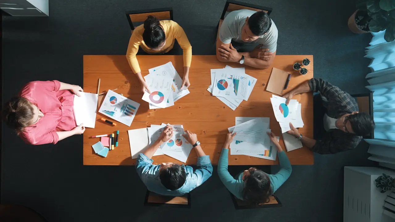 overhead view of a group of six people sitting at a table. they are discussing sheets of paper with various graphs and charts on them.