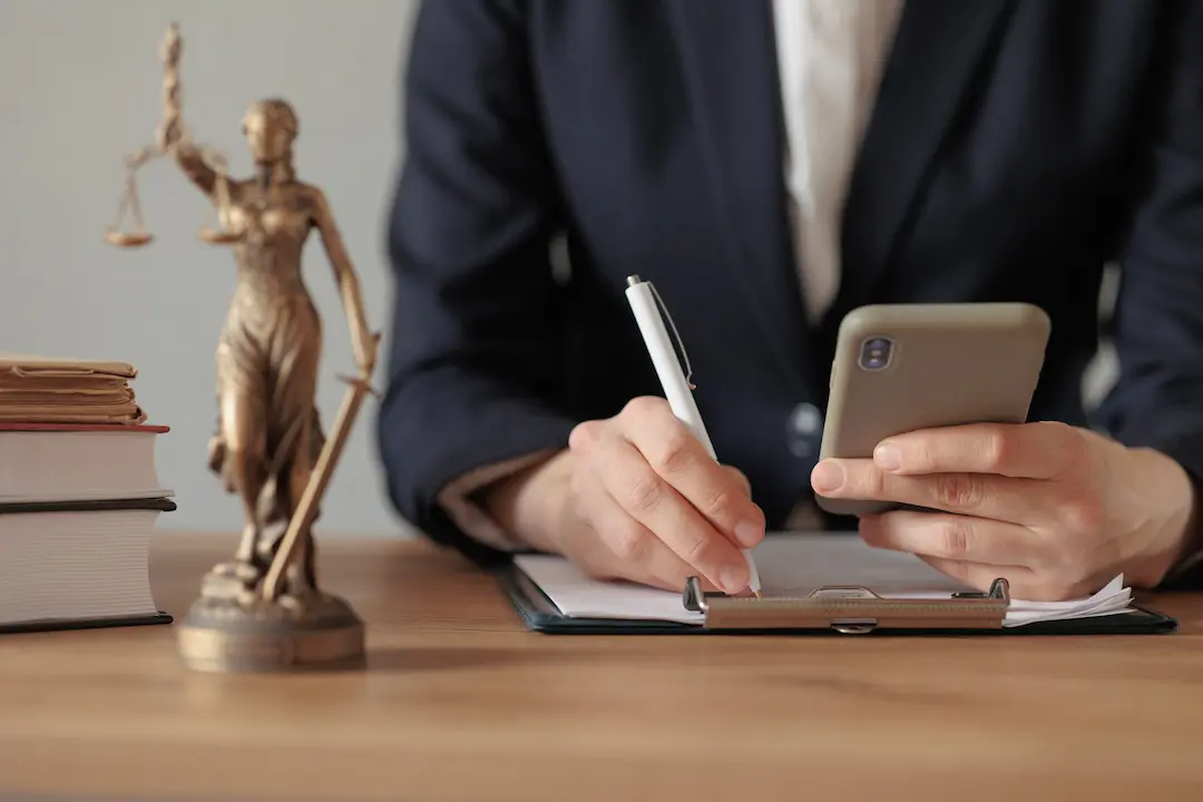 woman sitting at a table writing on a clipboard with one hand, holding a smartphone in the other. a small statue of justice holding the scales is to her right on the table.